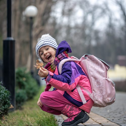 A little girl in a jacket, a hat with a backpack and a backpack examines the leaves outside, on the way to school or from school, the cold season.